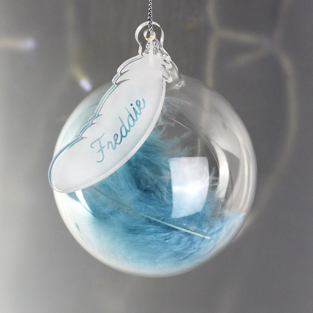 Decorative ornament with blue feathers inside a clear sphere and a name tag on a dark background