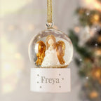 Decorative angel ornament in a clear ball with gold accents, hanging against a blurred Christmas tree background.