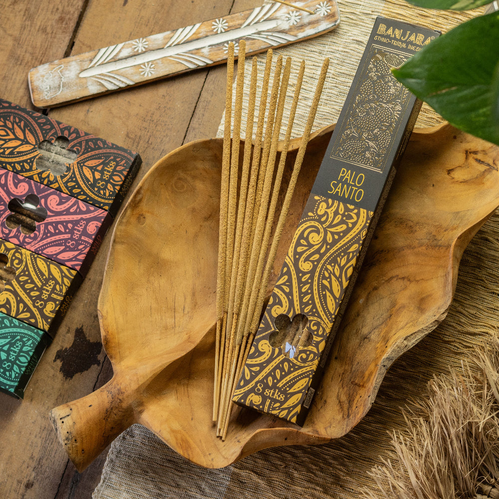A box of Banjar Palo Santo incense sticks sits on a carved wooden leaf dish, with several incense sticks leaning out. The scene is styled with a wooden spoon, a patterned box, and dried grass on a rustic wooden table.