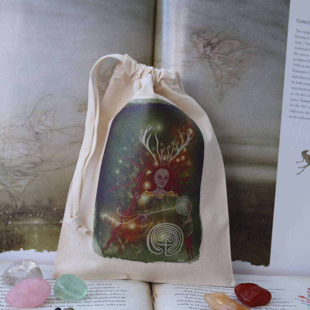 A beige fabric drawstring pouch featuring a colorful fantasy illustration of a fairy with antlers, displayed on open books with crystals and a beaded necklace around it.