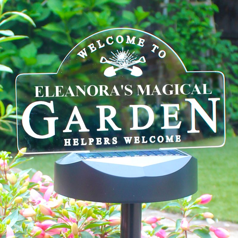 A clear acrylic garden sign reading 'Welcome to Eleanora's Magical Garden Helpers Welcome' mounted on a black solar-powered stake in a garden with green foliage and pink flowers.