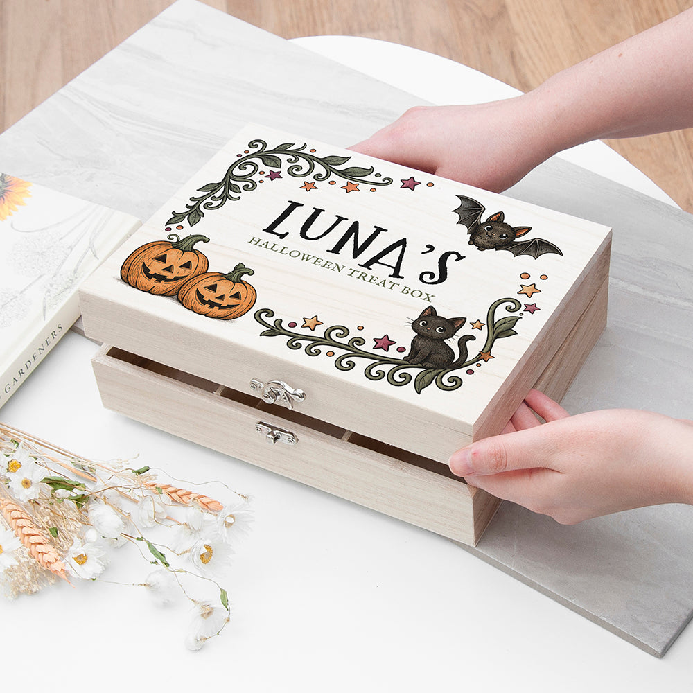 A wooden Halloween treat box with a sliding lid, decorated with illustrations of pumpkins, a black cat, a bat, and the text 'LUNA'S HALLOWEEN TREAT BOX'. Hands are holding the box on a light gray surface.