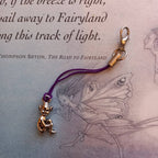 A gold fairy charm with a purple cord and lobster clasp, resting on a vintage book page with a fairy illustration and text.