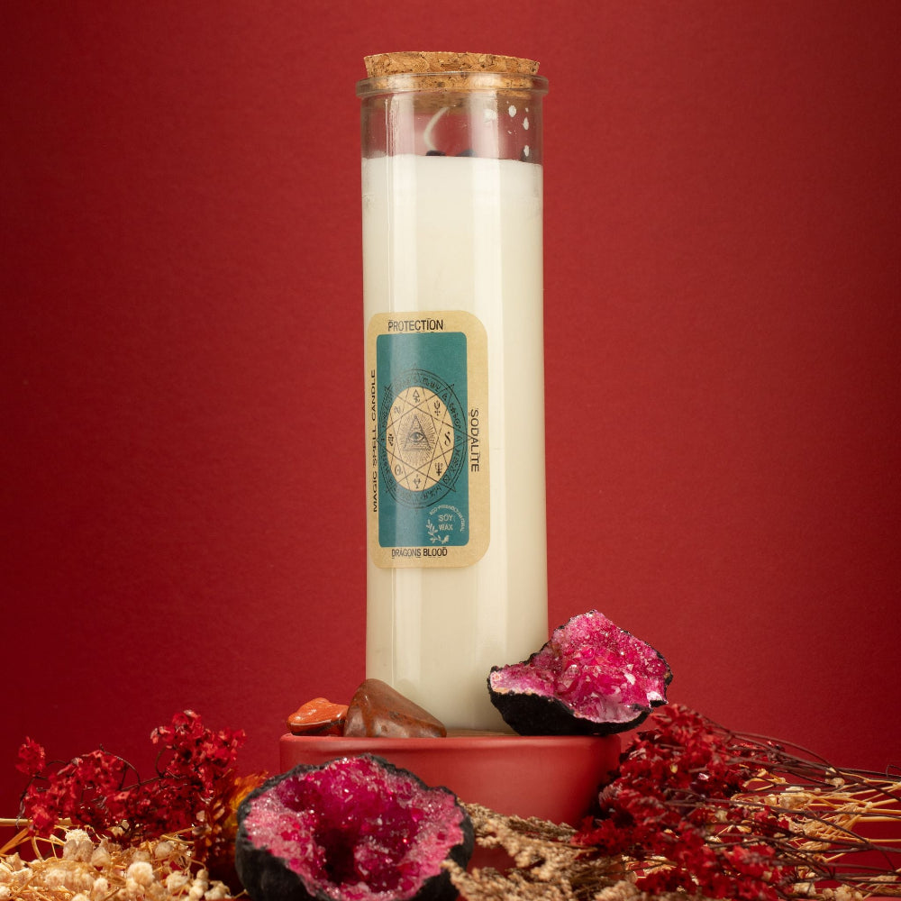A tall glass jar candle with a cork stopper and cream-colored wax stands on a terracotta base against a deep red background. The front label reads 'PROTECTION' 