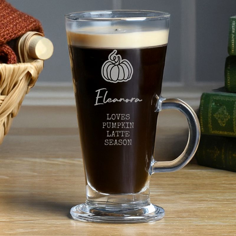 A clear glass latte mug filled with dark coffee, featuring a white pumpkin graphic and the text 'Eleanor LOVES PUMPKIN LATTE SEASON' on the front, set on a wooden tabletop with a basket and books in the background.