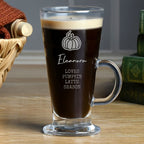A clear glass latte mug filled with dark coffee, featuring a white pumpkin graphic and the text 'Eleanor LOVES PUMPKIN LATTE SEASON' on the front, set on a wooden tabletop with a basket and books in the background.