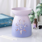 A lavender and light blue gradient ceramic oil burner with a sun and moon cutout, styled on a white surface with a reed diffuser stick, a small amber bottle, and a book in the background.