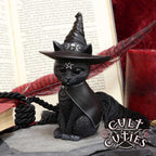 Black cat figurine wearing a witch hat with a pentagram symbol, sitting on a surface with an open book and red candle in the background.