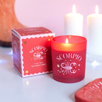 A red frosted glass scented candle labeled 'SCORPIO' stands next to its matching red and pink box on a light surface. Two blurred white candles and a warm-toned decorative object appear in the background, creating a cozy setting.
