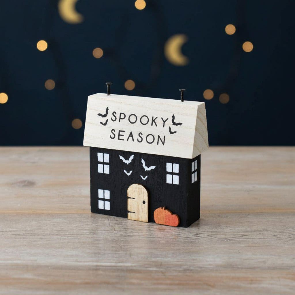 A small decorative house-shaped candle holder or ornament with a black body, light wood roof, white window details, a light wood door, and a small orange pumpkin at the base. The roof reads “SPOOKY SEASON” with bat silhouettes.