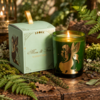 A green glass jar candle with a gold fairy illustration sits on a wooden table next to its pale mint-green box with a gold ribbon handle. The box is labeled 'Moss & Fern Scented Candles'.