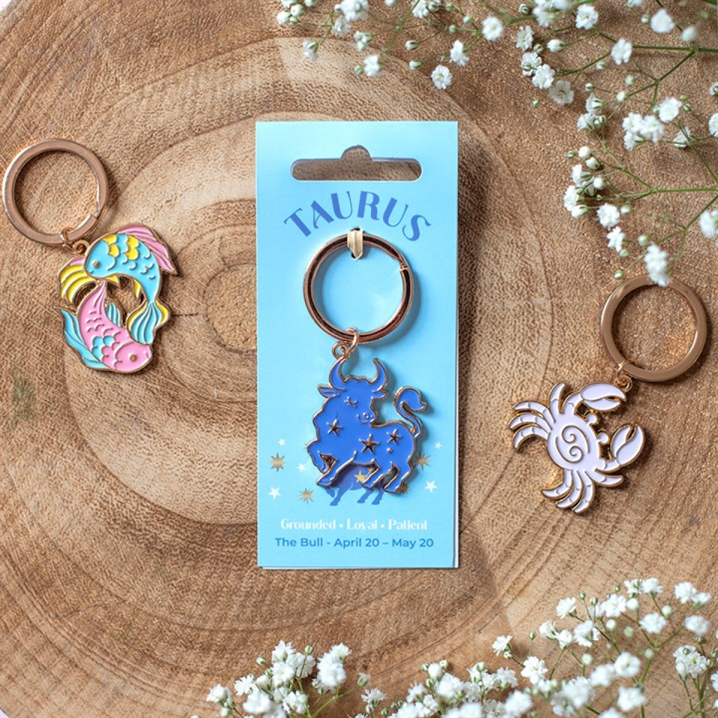 Three metal keychains displayed on a wooden surface with small white flowers. The center keychain is on a light blue card labeled TAURUS and shows a blue bull with stars; the left keychain is a colorful mermaid fish design, and the right keychain is a pink crab.