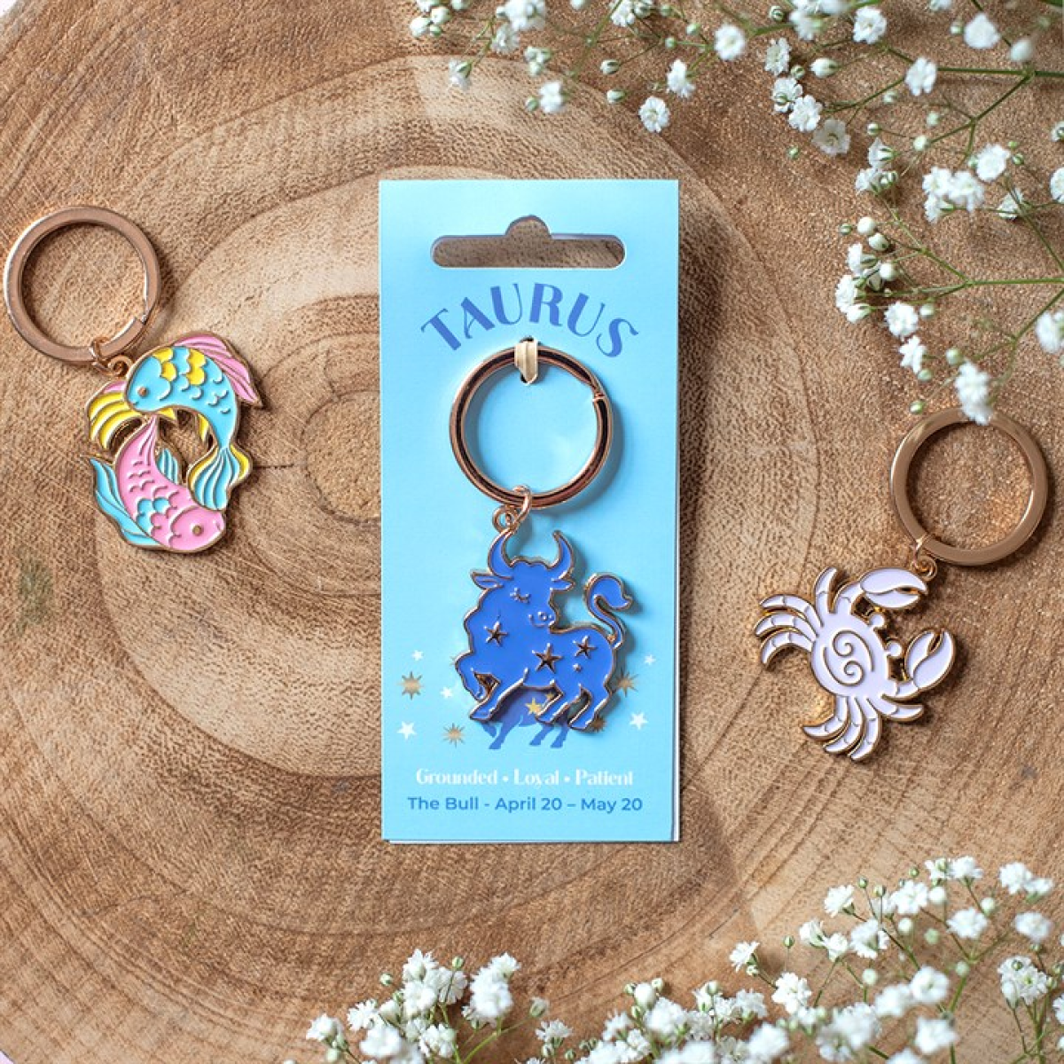 Three metal keychains displayed on a wooden surface with small white flowers. The center keychain is on a light blue card labeled TAURUS and shows a blue bull with stars; the left keychain is a colorful mermaid fish design, and the right keychain is a pink crab.