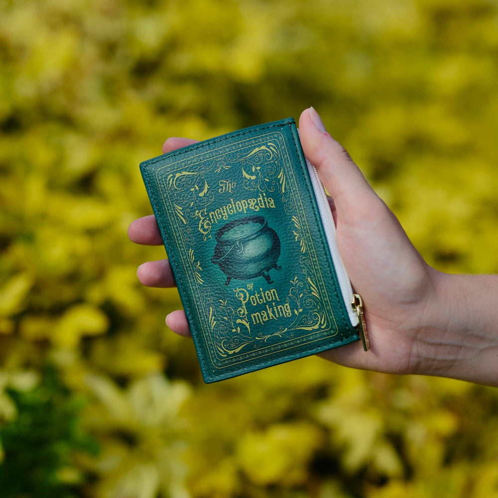 Hand holding a green book titled 'The Encyclopedia of Potion Making' against a blurred natural background