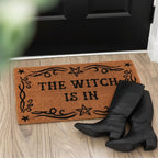Doormat with 'The Witch is In' text on a wooden floor, with black boots at the entrance.
