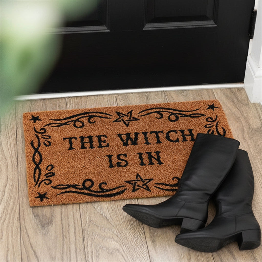 Doormat with 'The Witch is In' text on a wooden floor, with black boots at the entrance.