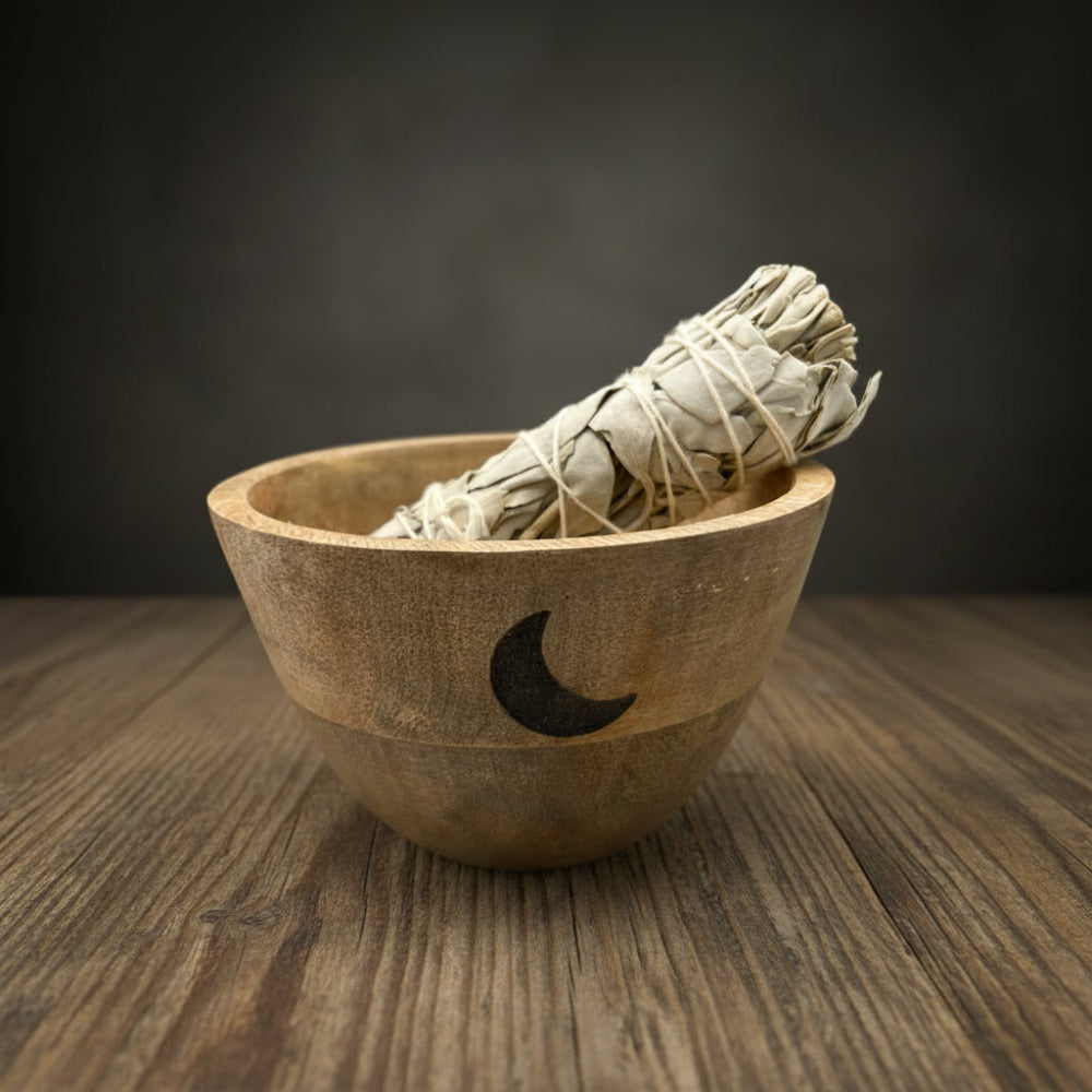 Wooden bowl with a crescent moon design containing a smudge stick, surrounded by greenery.
