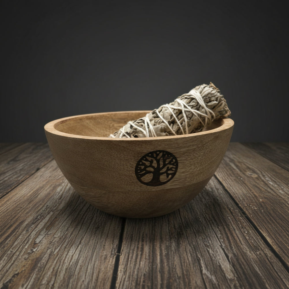 Wooden bowl with a tree design containing sage, surrounded by eucalyptus leaves on a neutral background.