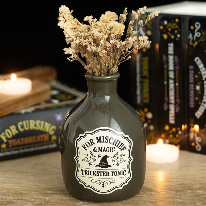 Decorative bottle labeled 'Trickster Tonic' with dried flowers, candles, and books in the background.