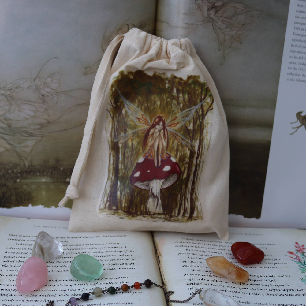 A cream-colored fabric drawstring pouch featuring a painted illustration of a fairy under red-and-white mushrooms, displayed on top of open books with crystals and a beaded necklace nearby.