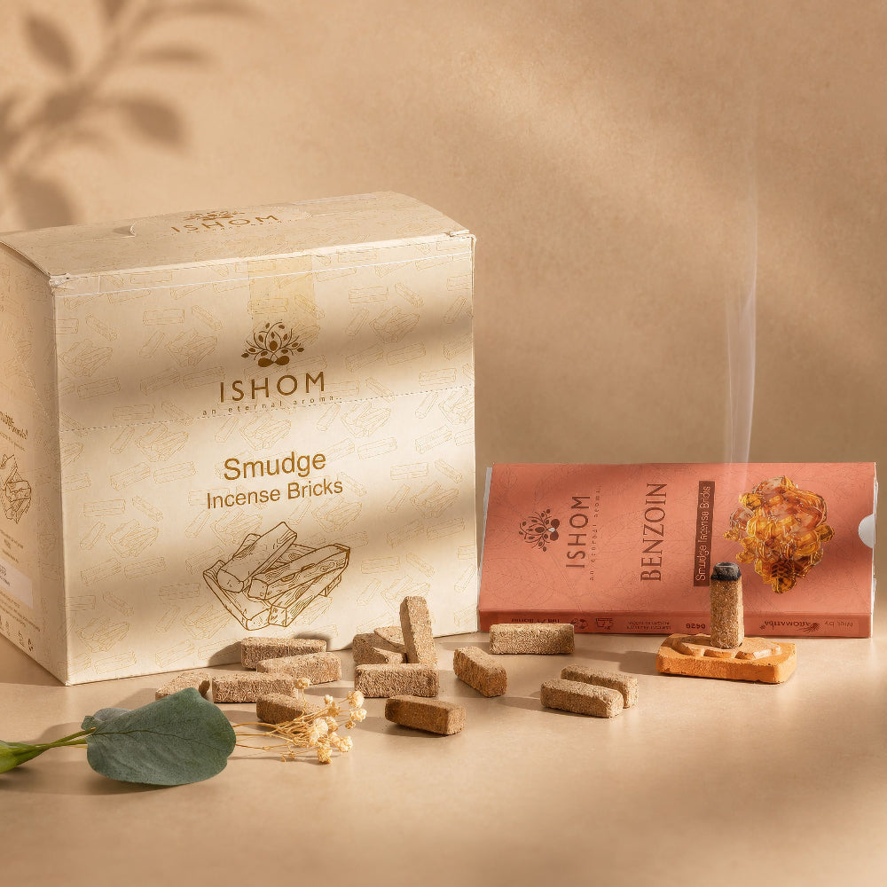 A styled product shot of Ishom Smudge Incense Bricks, showing a cream-colored product box, a pink benzoin box, several loose incense sticks, and one lit incense stick with visible smoke against a warm beige background.