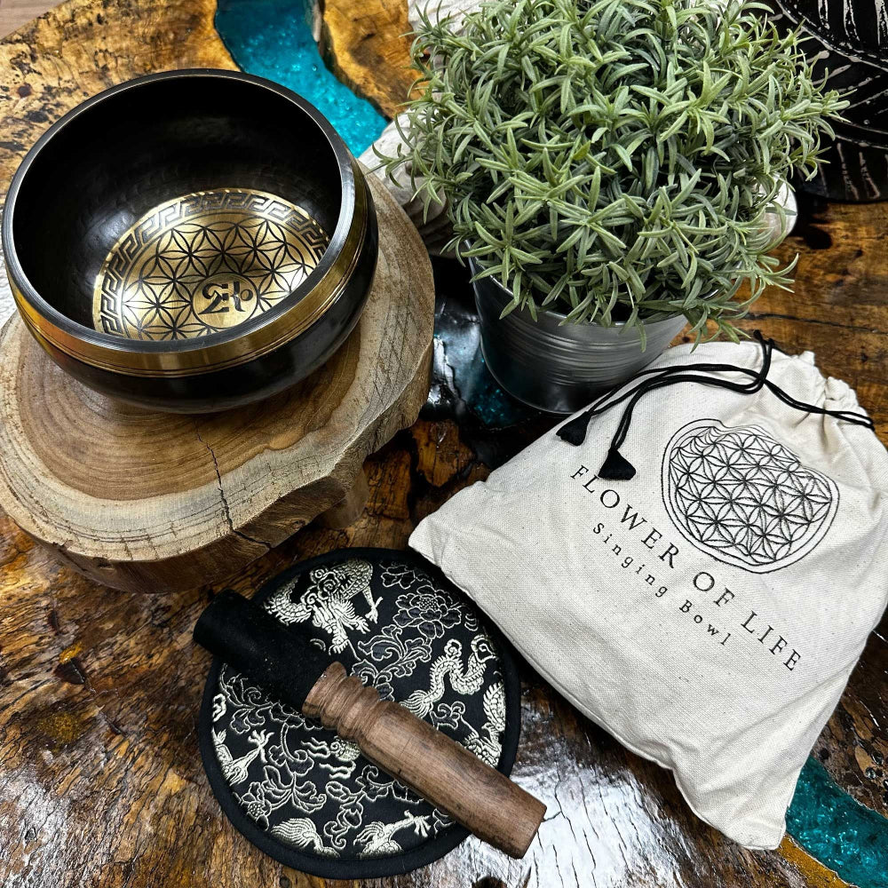 Singing bowl set with 'Flower of Life' design on a wooden surface with a plant and packaging.