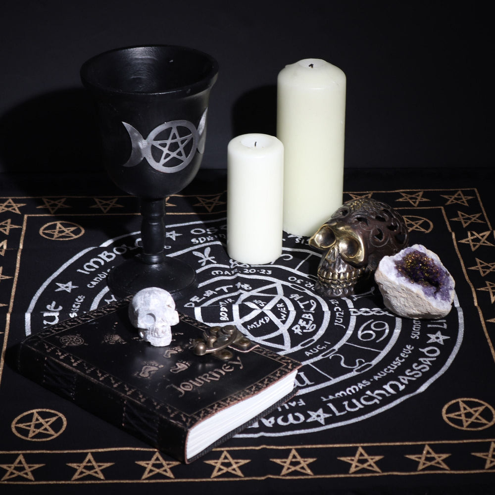 Witchcraft altar setup with black goblet, candles, skull, and crystal on a black cloth with white symbols.