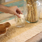 Hand placing a small candle in a glass jar on a wooden table.