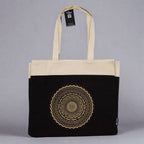 Tote bag with black bottom half and beige top half, featuring a mandala design. Eleanoras