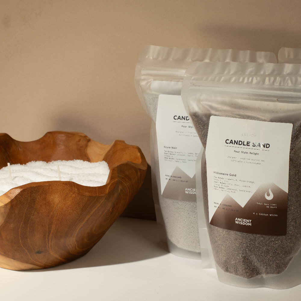 Two clear stand-up pouches of Candle Sand body scrub with white labels are shown on a tabletop beside a wooden bowl filled with white scrub, against a warm beige background.