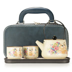 A tea set with a teapot, two cups, and a matching carrying case. The teapot and cups are cream-colored with a floral print featuring a cat, while the carrying case is dark blue-gray with a handle and zippered closure.