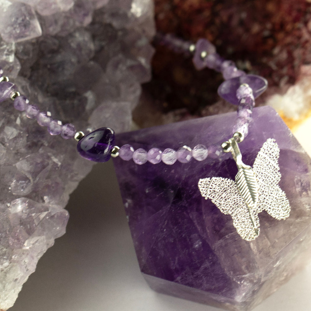 A beaded necklace with purple translucent beads and a silver butterfly pendant, displayed on purple crystal stones.