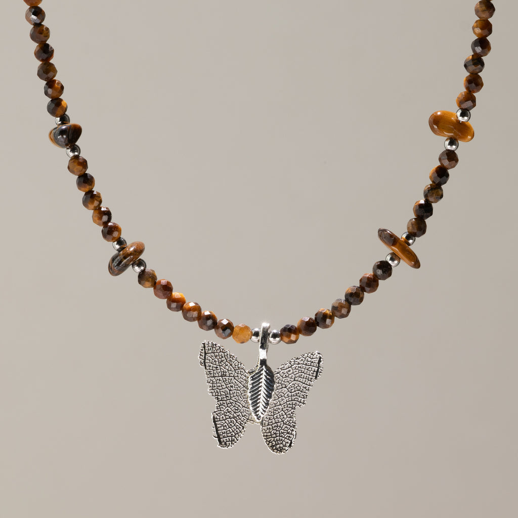A beaded necklace featuring brown and amber tiger's eye beads with a textured silver butterfly pendant, displayed against a solid warm gray background.