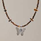 A beaded necklace featuring brown and amber tiger's eye beads with a textured silver butterfly pendant, displayed against a solid warm gray background.
