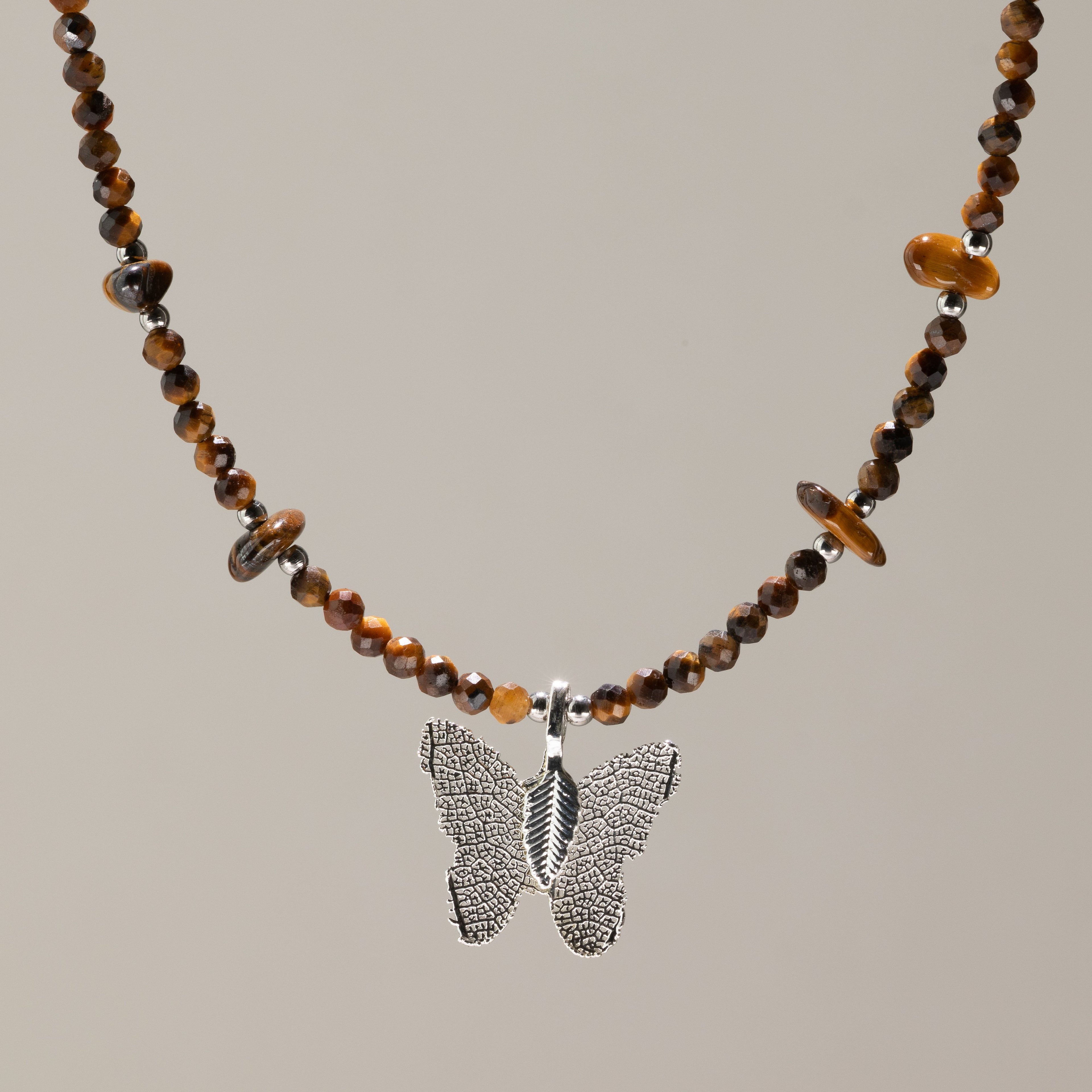 A beaded necklace featuring brown and amber tiger's eye beads with a textured silver butterfly pendant, displayed against a solid warm gray background.