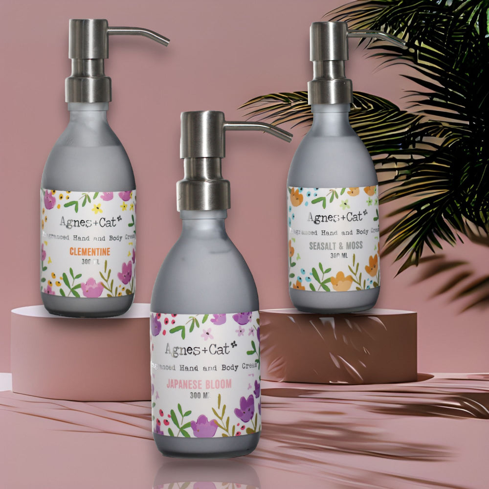 Three Agnes & Cat hand and body cream bottles with floral labels on a pink background.