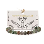 A beaded bracelet made of moss agate stones, displayed in front of its white paper packaging card. The card features gold and black text reading 'PETITE POWER BRACELET Moss Agate' with an illustration of hands holding a crystal.