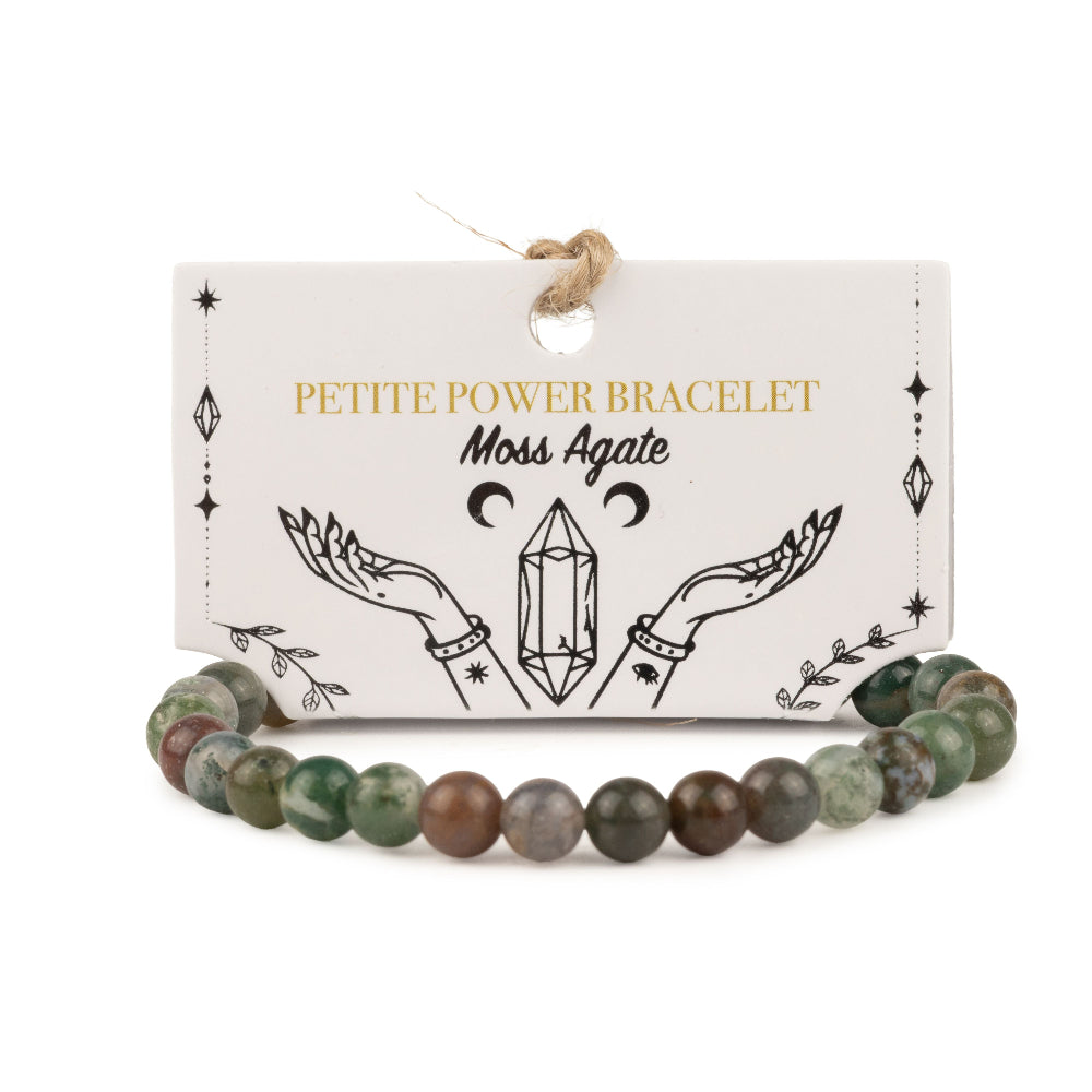 A beaded bracelet made of moss agate stones, displayed in front of its white paper packaging card. The card features gold and black text reading 'PETITE POWER BRACELET Moss Agate' with an illustration of hands holding a crystal.