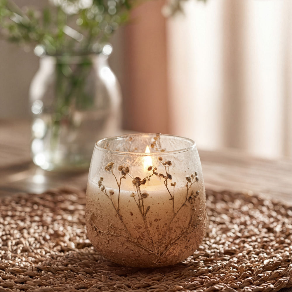 Candle in a glass holder with dried flowers on a textured surface, with a blurred vase of greenery in the background.
