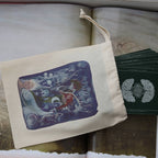 A cream canvas drawstring pouch with a colorful fantasy illustration, resting on an open book next to a fanned deck of dark green cards with silver circular logos.