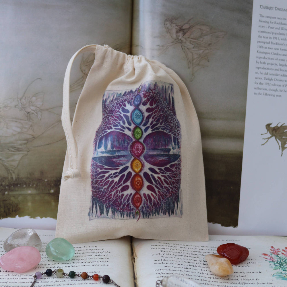 A beige drawstring pouch featuring a colorful chakra tree graphic, displayed on an open book with crystals and a pendant nearby.