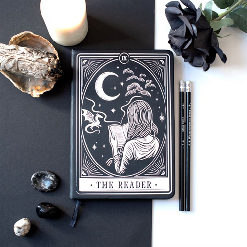 A tarot-style notebook or journal with the card 'The Reader' displayed on a split black-and-white background, surrounded by a candle, dried flower, black rose, pencils, and small stones.
