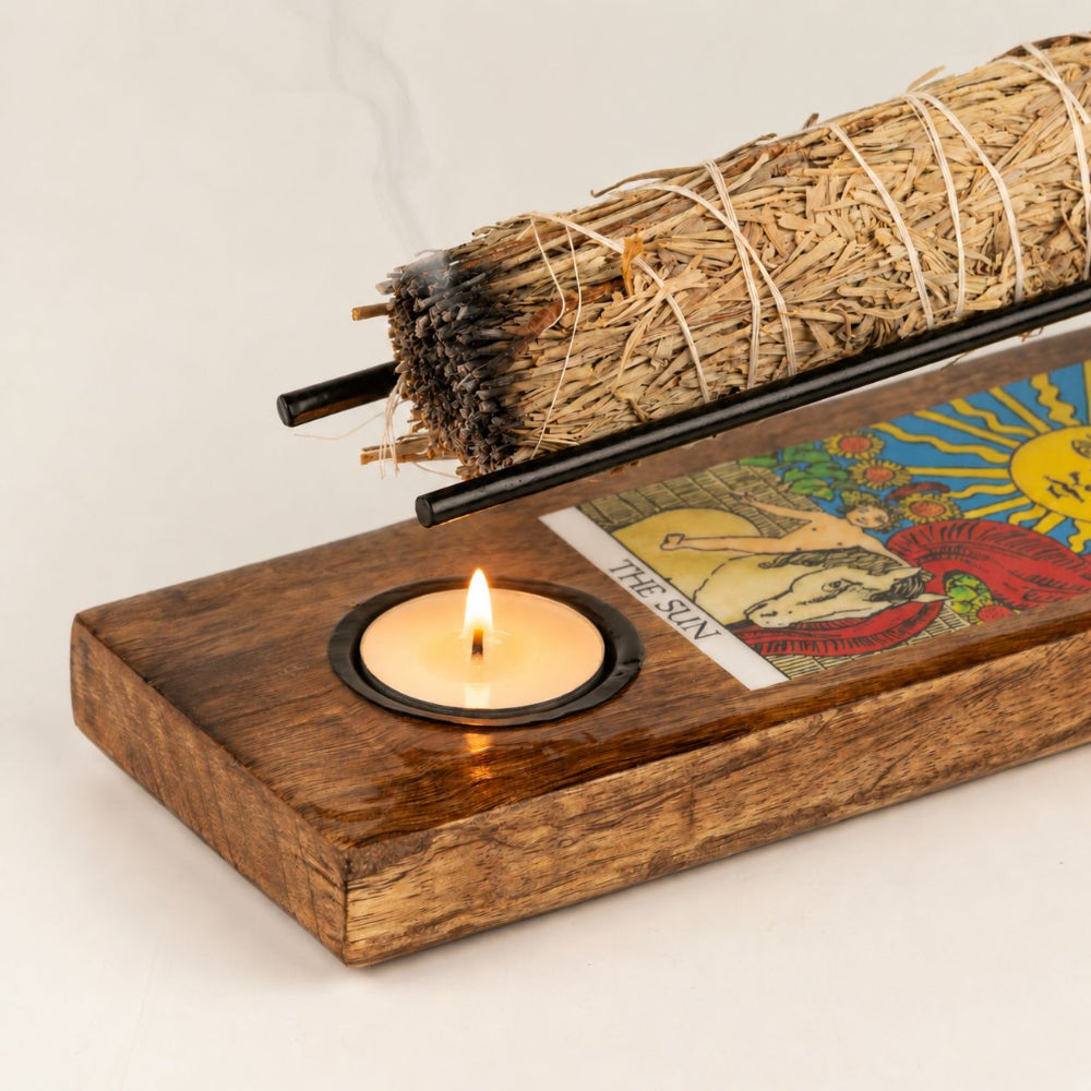 A wooden altar-style holder with a built-in tea light candle and a colorful tarot card panel, shown with a bundle of dried grass and a black rod resting across it on a light neutral background.