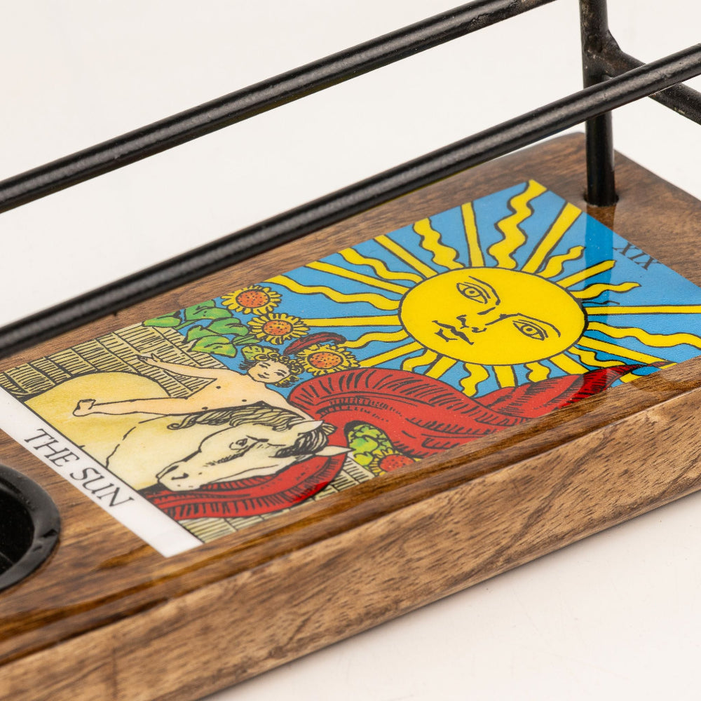 A close-up of a wooden tray or coaster featuring a colorful printed card design with the Sun of Wands tarot card artwork, alongside a black metal stand on a white background.